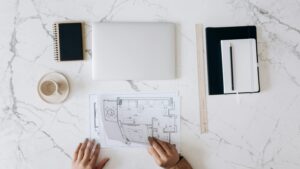 pexels-photo-6894105-6894105 Top view of an architect's desk featuring blueprints, laptop, notebook, and coffee cup on a marble surface.